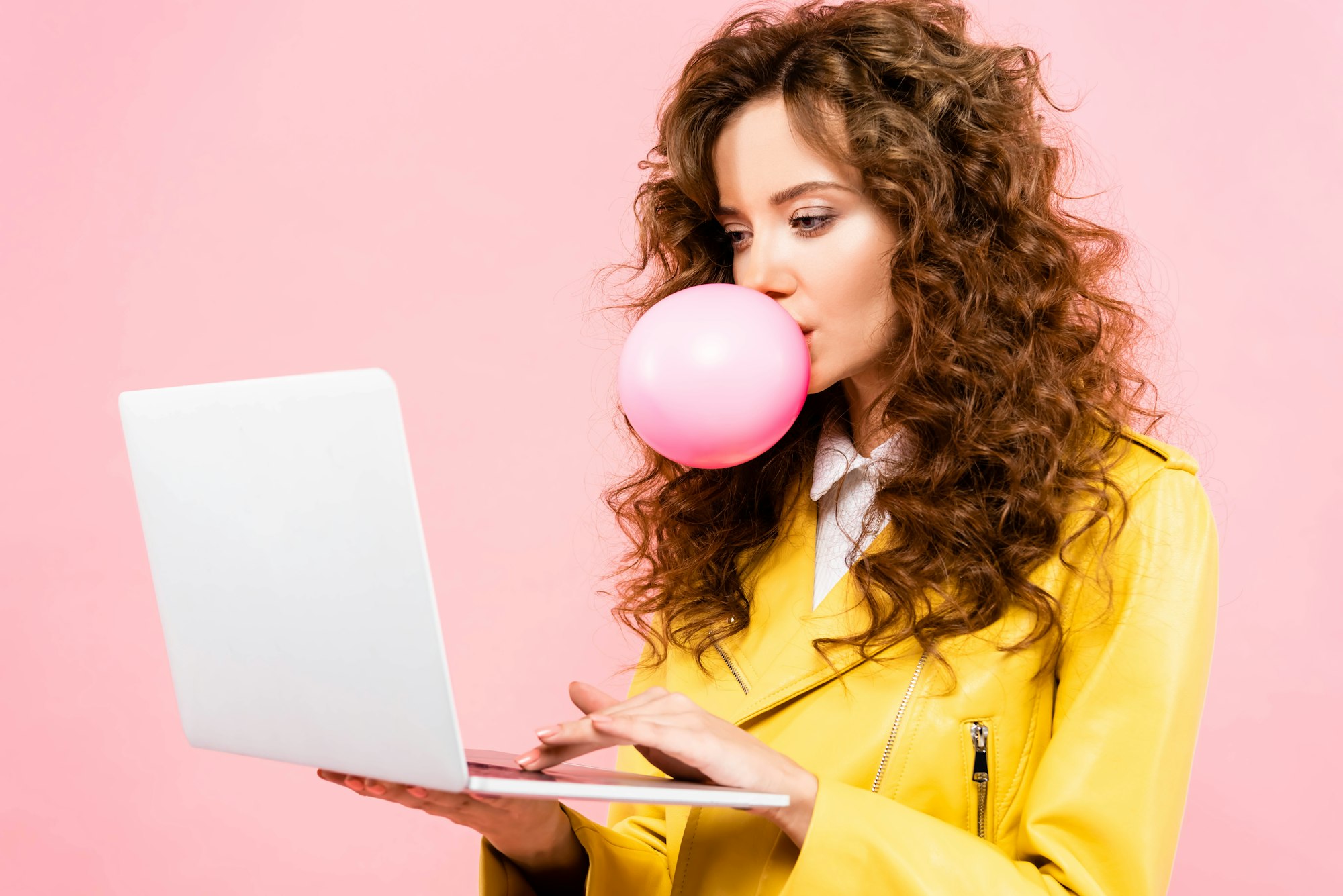happy curly woman with bubble of chewing gum using laptop, isolated on pink